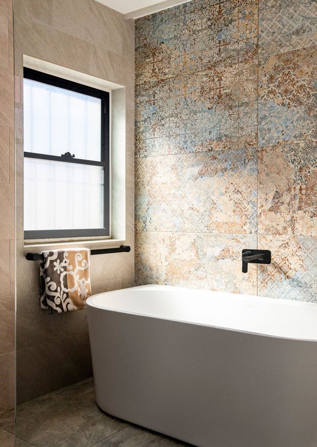 A close-up view of a refined bathroom featuring a freestanding white bathtub, a black-framed window, dark tapware and towel rail, large neutral wall tiles, and a prominent blue, beige and brown patterned feature-tile wall. The space is bright with a warm, textured and luxurious feel.