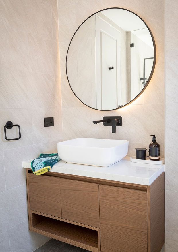 A bright contemporary bathroom vanity area with a wall-mounted timber vanity, white vessel basin, pale large-format tiled walls, matte black wall-mounted tapware, and a round backlit mirror. A black towel ring, electrical outlet, soap dispenser, and small tray are also visible.