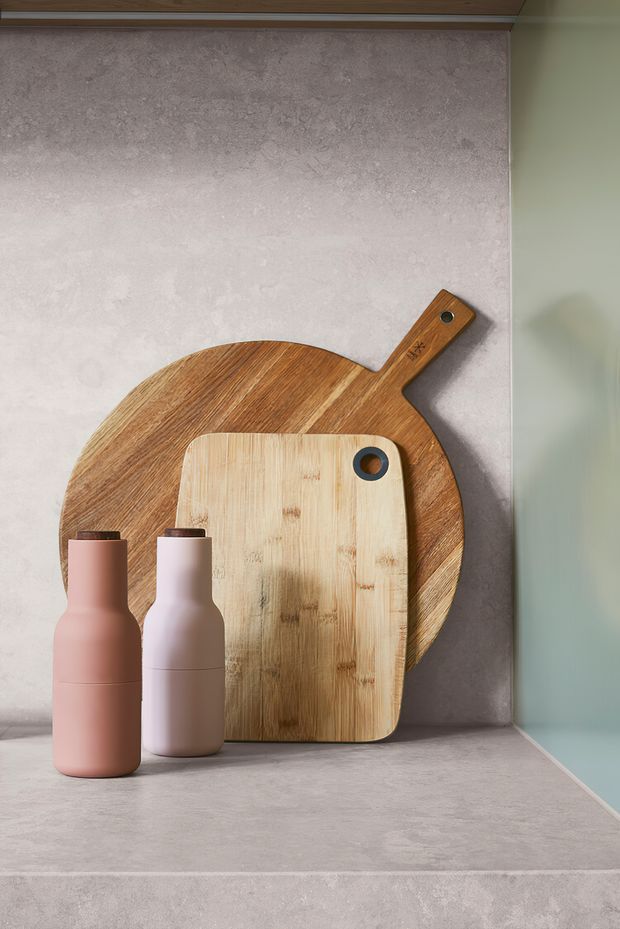 A styled kitchen benchtop vignette with two pastel bottles and overlapping wooden serving and chopping boards. The scene features a pale stone-look benchtop and matching textured splashback, a muted sage-green side wall, and warm timber cabinetry or trim at the top.