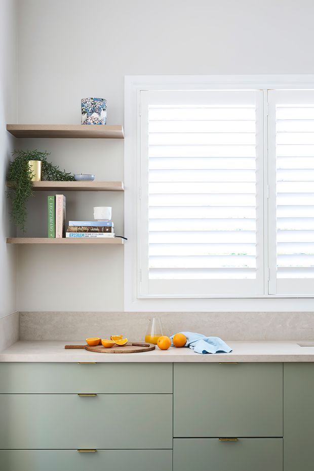 Bright, compact kitchen vignette featuring muted sage-green lower cabinetry with slim brass pulls, a pale stone or composite benchtop and backsplash, open timber shelves with books and decorative objects, and a large white shuttered window. Citrus fruit, a cutting board, glass jug, and blue cloth add a lived-in, fresh feel.