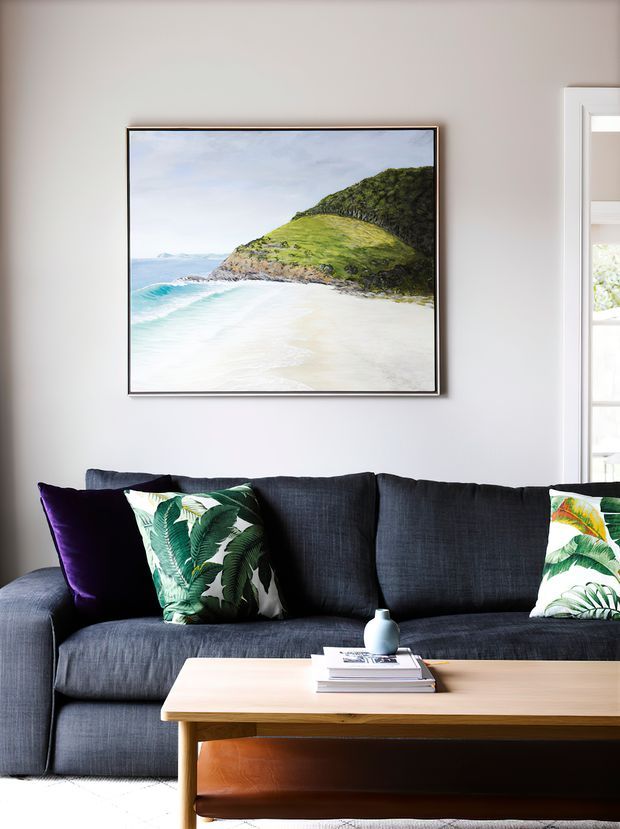 A styled living room with a dark charcoal sectional sofa, tropical botanical cushions, and a light timber coffee table with a leather sling shelf. A framed beach landscape artwork hangs on a pale wall, with a bright adjoining space visible through the doorway.