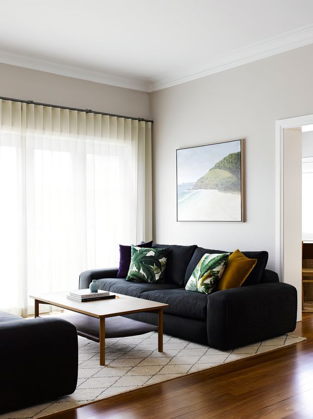 Bright, refined living room with a large dark charcoal sectional sofa, patterned botanical and mustard cushions, a simple light timber coffee table, and a pale geometric area rug. The room has warm timber flooring, soft beige walls, sheer vertical blinds, white decorative ceiling cornices, and a framed coastal landscape print. An open doorway provides a glimpse into an adjacent room.