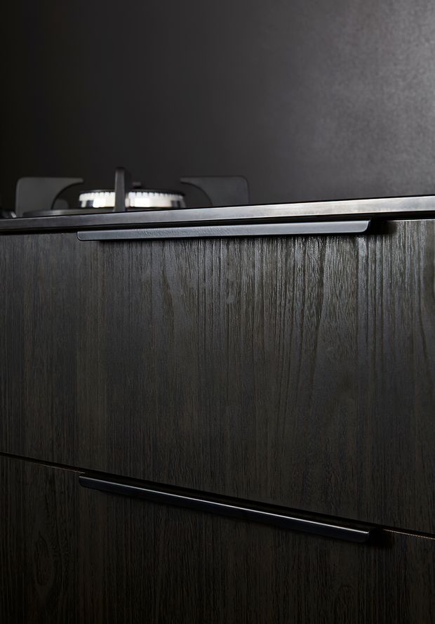 Close-up detail of dark vertically grained timber-look cabinetry with sleek black or dark metal recessed handles. A partial gas cooktop is visible behind the cabinet front, suggesting a dark modern kitchen fit-out. The image is tightly framed and moody, focused on the cabinetry finish and hardware.