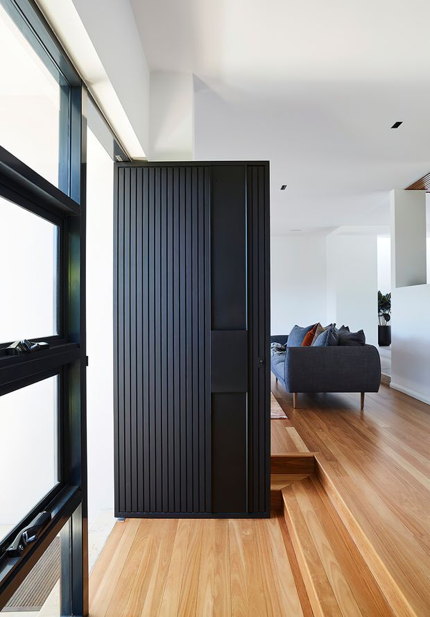 Bright contemporary interior featuring a dramatic black vertical-slat pivot door or screen beside full-height black-framed glazing. Warm timber flooring continues through a small stepped level change into an open living area with a dark grey sofa, while white walls and ceiling create a clean, minimal setting.