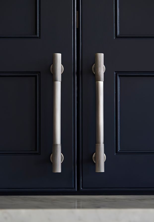 Close-up detail of dark navy or charcoal paneled cabinetry with two long vertical decorative handles. The handles combine ribbed metallic end caps and pale cylindrical central sections, with a light stone or marble-like surface visible below. The image emphasizes refined, high-contrast joinery hardware and moulded cabinet detailing.