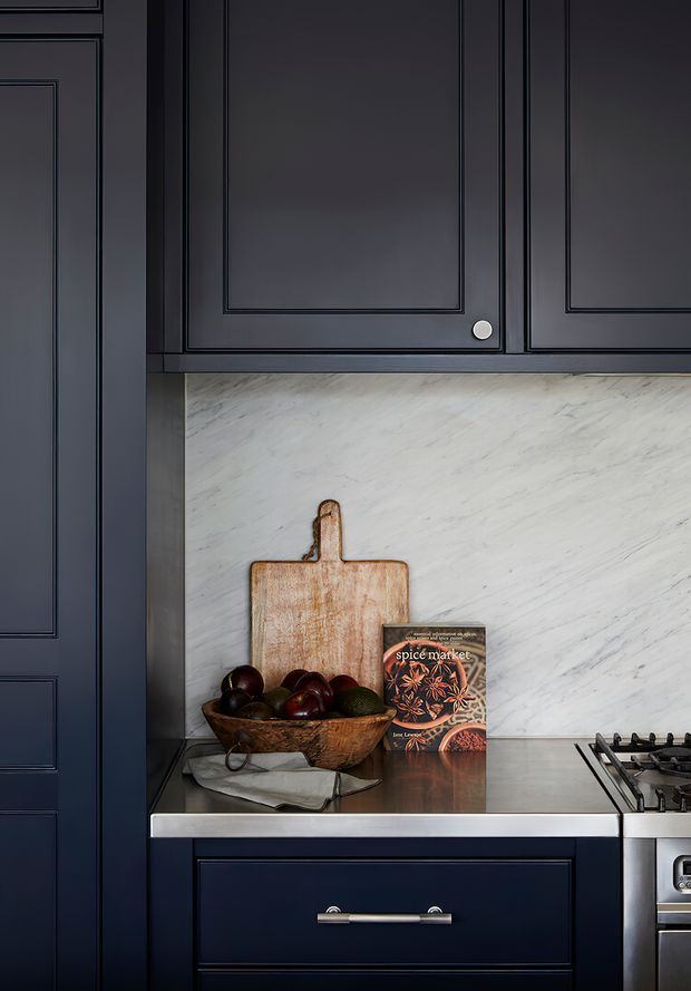Close-up view of a refined dark navy kitchen, featuring raised-panel cabinetry with brushed metal hardware, a pale veined stone backsplash, dark reflective benchtops, a gas cooktop, and a lower drawer unit. A timber chopping board, fruit bowl, and cookbook add warmth to the composition.