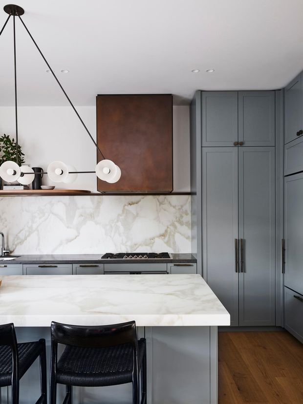 A refined kitchen with full-height muted grey-blue panelled cabinetry, a large white marble-look island and matching splashback, warm timber flooring, and black woven leather bar stools. A sculptural linear pendant with round white globes hangs above the island, while a dark bronze feature cabinet adds contrast above the cooktop.