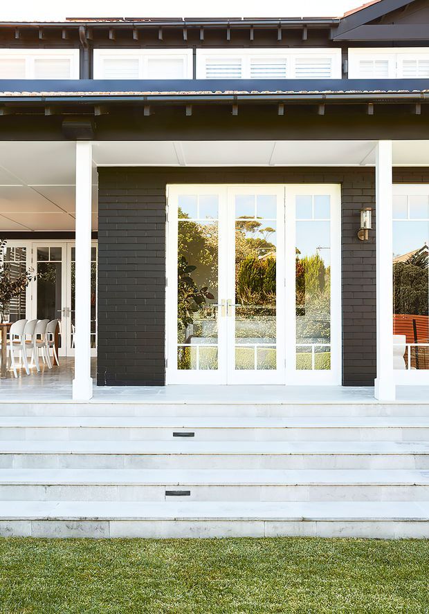 Front exterior view of a contemporary residential home with a covered entry porch, broad white steps, white-framed French doors, dark charcoal brickwork, white columns and trim, and a dark pitched roof. A dining area is visible through the left glazing, with landscaped garden surroundings reflected in the doors.