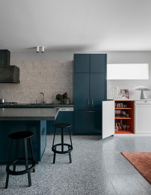 Contemporary kitchen interior with a dark blue island and tall pantry cabinetry, pale textured square-tile splashback, dark stone benchtops, integrated appliances, and black bar stools. A white built-in sideboard includes an open wine-glass cabinet with warm timber-toned interior. The space has a light grey terrazzo-like floor, white ceiling, and a large horizontal window.
