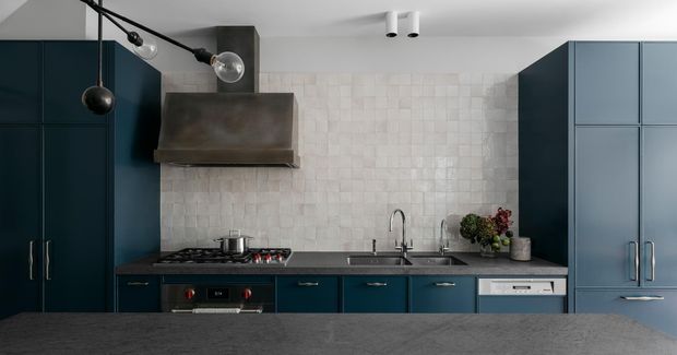 Contemporary galley-style kitchen with dark blue floor-to-ceiling cabinetry, a dark stone benchtop, pale textured square-tile splashback, integrated stainless-steel appliances, and a sculptural black-and-glass pendant light. The compact layout is viewed from across a foreground island or counter.