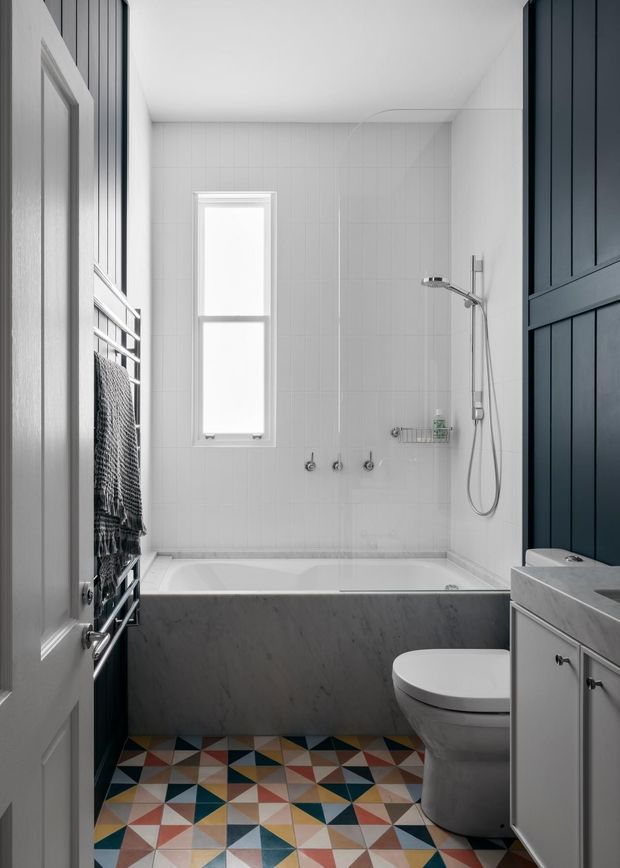 A compact, bright bathroom viewed through its doorway, featuring a white tiled bath and shower enclosure, a grey stone-look bath panel, white toilet and vanity, dark blue vertical wall panelling, a narrow window, heated towel rail, and a colourful geometric patterned floor tile.