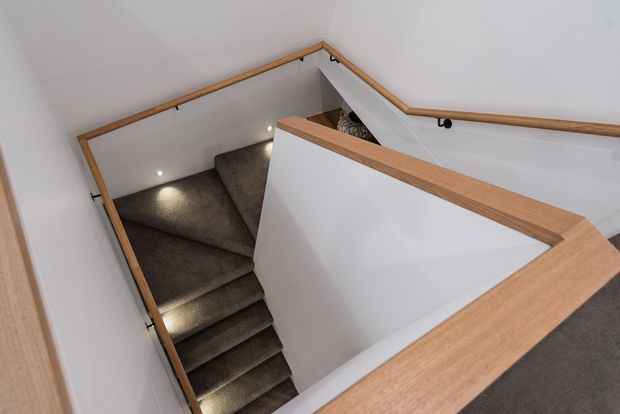 A compact stairway viewed from above, with dark carpeted treads and landing, white walls, recessed step lighting, and warm timber handrails and balustrade trim. The stair turns at a small intermediate landing within a narrow stairwell.