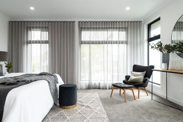 Bright, refined bedroom with a white upholstered bed, grey bedding, full-height grey curtains, large windows, a patterned area rug, and a mid-century-inspired charcoal lounge chair with matching ottoman. A slim console with decorative objects sits beneath a round mirror, while recessed ceiling lights provide even illumination.