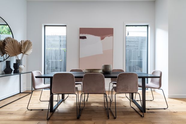 Bright contemporary dining room with a long dark dining table surrounded by upholstered blush-toned chairs. The room has pale walls, wide light timber flooring, tall narrow windows, a large abstract artwork, and a minimalist sideboard with sculptural dried botanical decor.