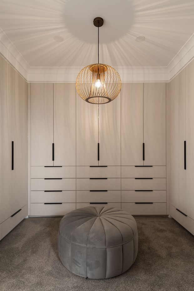 A refined walk-in wardrobe with full-height pale timber or wood-look built-in cabinetry, integrated drawers and slim black handles. A central round upholstered ottoman sits on a soft grey carpet, beneath a sculptural brass wire pendant light. White decorative ceiling cornices and restrained detailing create a luxurious, calm dressing-room atmosphere.