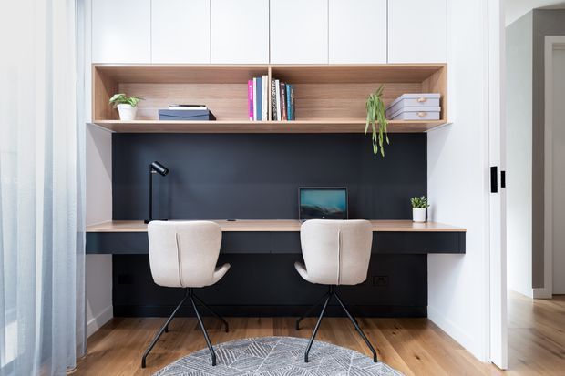 A bright built-in home office with a long timber desk spanning a dark feature wall, two upholstered swivel chairs, integrated white overhead cabinetry, an open timber display shelf with books and plants, and a small desk lamp. Timber flooring, light curtains, and a patterned rug create a clean, contemporary workspace.