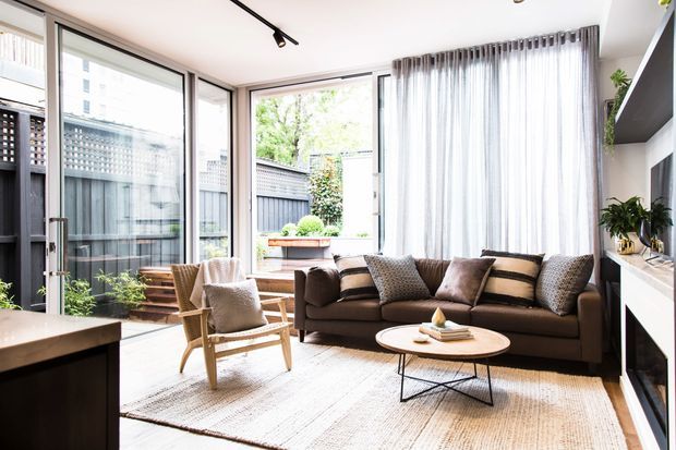 Bright contemporary living room with a dark sectional sofa, timber-framed lounge chair, round coffee table, woven rug, sheer curtains, and floor-to-ceiling glazing opening toward a small planted outdoor area. A built-in media wall and shelving are visible at right, with a compact dining or kitchen surface in the foreground.