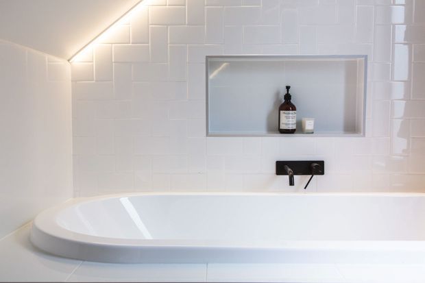 A clean, contemporary bathroom featuring a white freestanding bathtub, glossy white rectangular wall tiles, a recessed wall niche with bath products, and a matte black wall-mounted tap fitting. Soft concealed lighting highlights the tiled wall and niche.