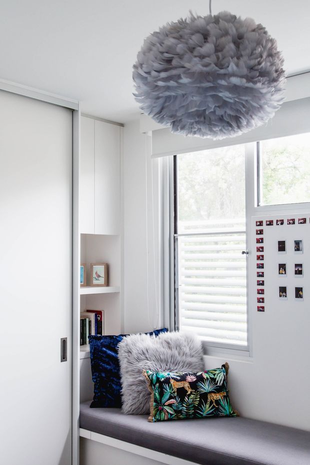 Bright, compact built-in window seat in a white room, with grey upholstered bench seating, colourful patterned cushions, a small bookshelf niche, framed photographs, and a large sculptural feather-like pendant light. A sliding white door and louvred window provide clean built-in architectural detailing.
