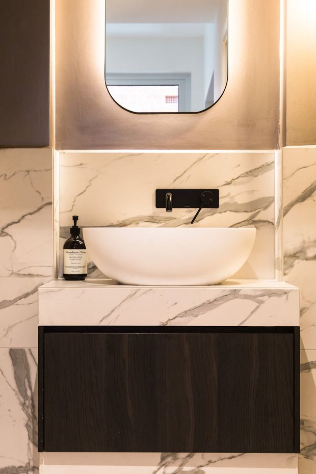 Close-up of a refined bathroom vanity featuring a white vessel basin, black wall-mounted tapware, dark timber vanity cabinetry, and white marble-look wall and benchtop surfaces with pronounced grey veining. A rounded-rectangle mirror with integrated warm lighting is mounted above.