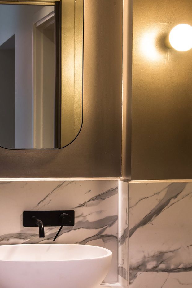Close-up of a refined bathroom vanity featuring a white vessel basin, matte black wall-mounted tapware, dramatic grey-veined marble wall tiling, a rounded-edge mirror, and warm brass-toned walls with a spherical wall light.