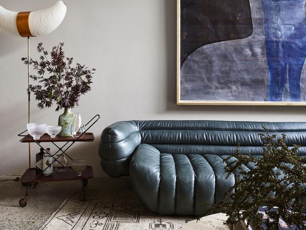 A stylish living room vignette featuring a sculptural dark blue-grey leather sofa, a vintage bar cart with ceramics and a vase of foliage, a curved floor lamp, patterned rug, and a large abstract artwork in muted blue, charcoal, and grey tones. The space has a refined retro character with strong mid-century influences and dramatic sculptural furniture.