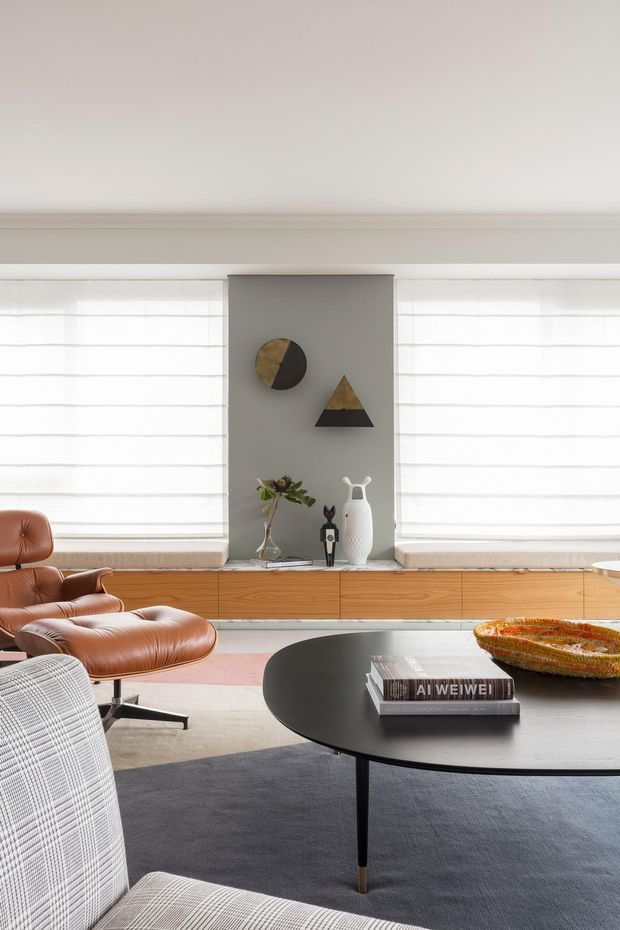 A refined living room with a low, long timber sideboard beneath broad windows fitted with white sheer Roman blinds. A grey feature panel displays geometric wall art and sculptural objects. The room is furnished with a cognac leather lounge chair and ottoman, a light patterned armchair, and a large black oval coffee table layered over a dark rug and pale flooring. The restrained palette and clean lines create a sophisticated mid-century-inspired interior.
