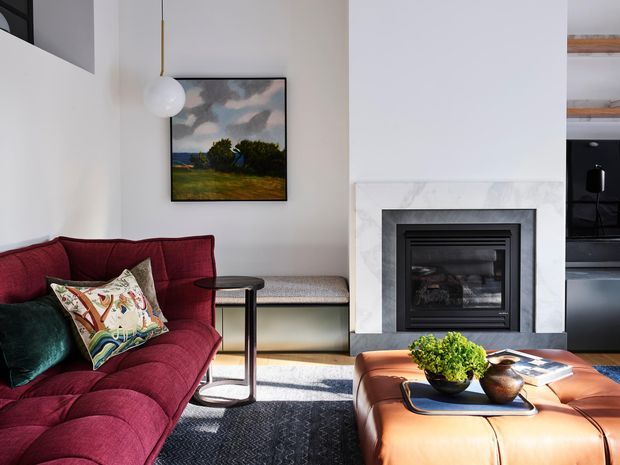 A refined living room with a deep burgundy sectional sofa, patterned and teal cushions, a low tan leather ottoman coffee table, and a black textured rug. A marble-framed fireplace forms the central architectural feature, with a small upholstered bench beneath an artwork and a sculptural pendant light. The room opens toward a dark-toned kitchen or joinery area at right and receives strong natural light.