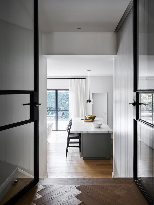 View through black-framed glazed doors into a bright, refined kitchen and dining area. The space features white vertical wall panelling, a dark kitchen island with a white marble-look benchtop, black bar stools, a linear pendant light, pale timber flooring, and full-height glazing with sheer curtains. The foreground has dark herringbone timber flooring and matching black-framed doors.