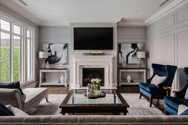 Elegant formal living room with grey paneled walls, a white classical fireplace mantel, wall-mounted television, large windows, symmetrical console styling, a glass-top coffee table, a neutral sectional sofa, and deep navy wingback chairs. The room combines traditional architectural detailing with refined contemporary furnishings.