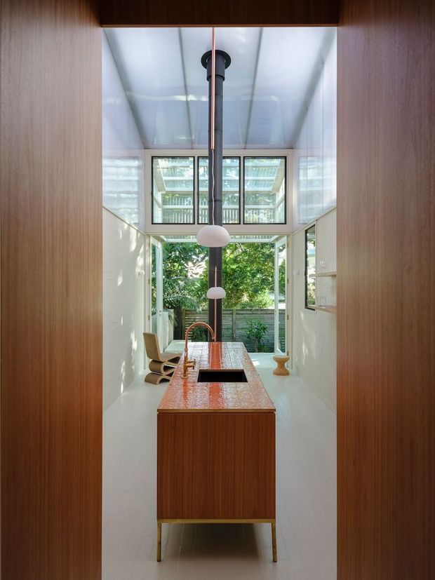 A narrow, double-height kitchen viewed through a timber-lined opening. The space features a central timber island with a glossy reddish patterned benchtop and integrated dark sink, white walls and floor, full-height glazing, and a tall black pendant light with globe shades. A small dining or sitting area and lush greenery are visible beyond the glazed doors.