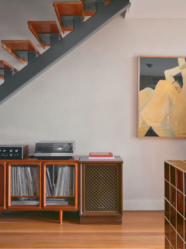 Interior detail of a stylish living room featuring a mid-century-inspired timber vinyl record cabinet and speaker, turntable, record collection, and framed artwork. A dark exposed stair stringer with timber treads crosses the upper left, while a glossy dark tiled or gridded cabinet is partially visible at right. The room has warm timber flooring, a pale neutral wall, and carefully composed retro audio furnishings.