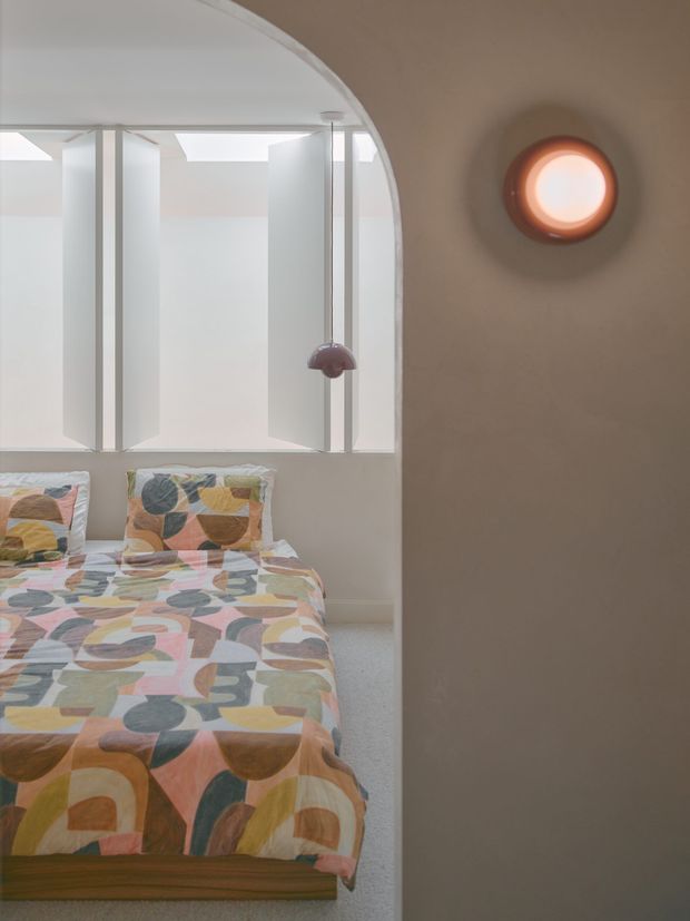 A compact, softly lit bedroom viewed through a curved opening. The room contains a bed with a colourful geometric-patterned duvet and pillows, pale walls, a broad multi-pane window with translucent privacy panels, and a small spherical pendant light. A rounded wall light glows on the warm taupe wall in the foreground. The overall interior is playful, retro-inspired, and muted in tone.