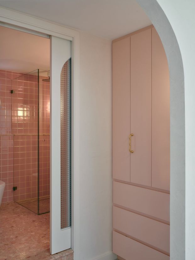 A compact pink-toned bathroom viewed through an arched opening, with a glass-enclosed shower lined in glossy square pink tiles, a terrazzo-like pink floor, and a full-height built-in blush-pink vanity or storage cabinet with drawers and a small brass handle. A white-framed glazed door and patterned translucent panel separate the shower area from the main space.