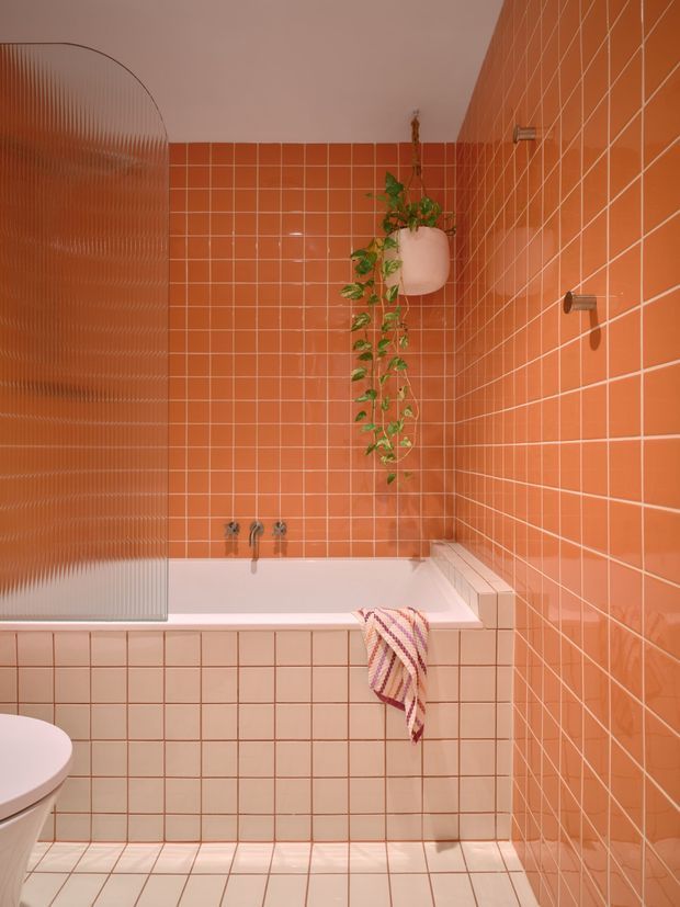 A compact, vividly styled bathroom with a built-in bathtub enclosed by a translucent ribbed glass screen. Coral-orange square tiles cover the walls, bath surround, and floor, with pale grout lines creating a strong grid pattern. A white toilet is partially visible, along with wall-mounted bath fixtures, a hanging white planter with trailing greenery, and a striped towel draped over the tub. The space has a playful contemporary feel and warm, saturated colour palette.