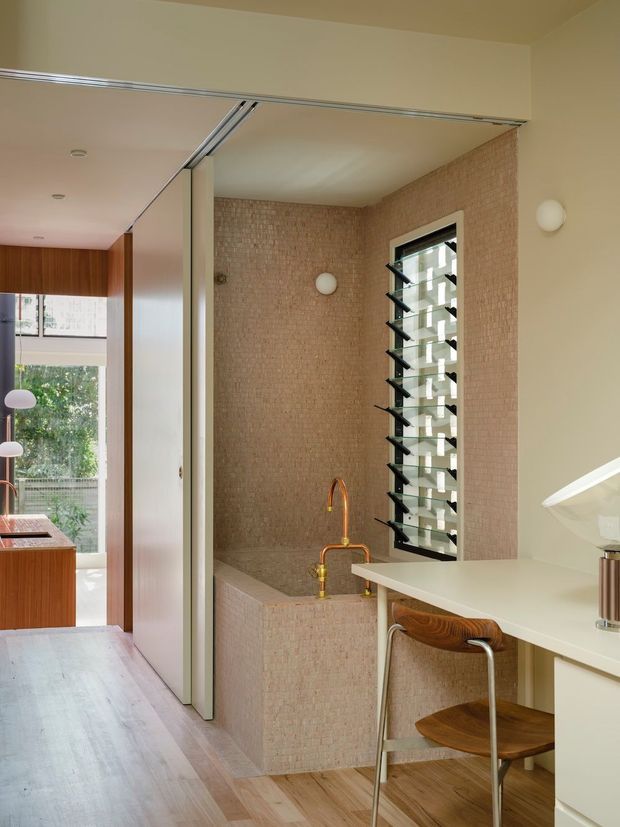 A compact contemporary kitchen and utility area with a pale stone or solid-surface island benchtop, integrated cabinetry, timber-fronted cabinetry in the background, and a built-in tiled or textured masonry sink/bath-like basin zone. The walls around the basin are clad in small warm beige tiles or textured brick, with a tall black-framed louvred window providing ventilation and daylight. A timber-and-metal bar stool sits at the island, and a sliding partition separates the kitchen from the adjoining glazed living or dining area.
