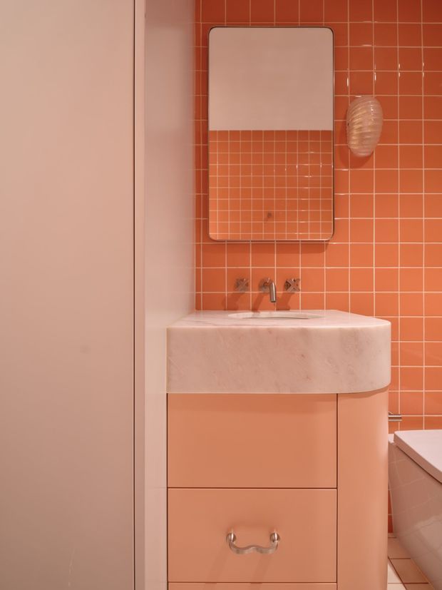 A compact, highly coordinated pink and terracotta bathroom featuring a rounded stone or solid-surface vanity basin with integrated counter, peach-pink cabinetry, glossy terracotta square-tile walls, a large rounded-rectangle mirror, and chrome tapware. A partial bathtub is visible at right. The space has a playful retro-inspired colour scheme and strong tonal cohesion.