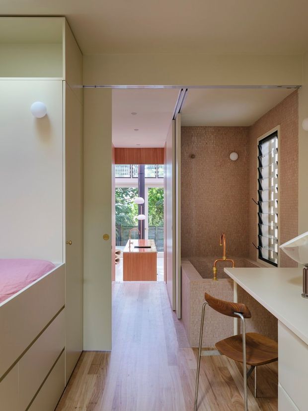 A compact contemporary bathroom viewed from an adjoining bedroom or dressing area, with a freestanding bathtub clad in small textured beige tiles, a white vanity with integrated basin, timber flooring, a louvred window, and warm timber cabinetry visible beyond the hallway. A partial bed is visible at left, confirming the bathroom connects directly to the bedroom.
