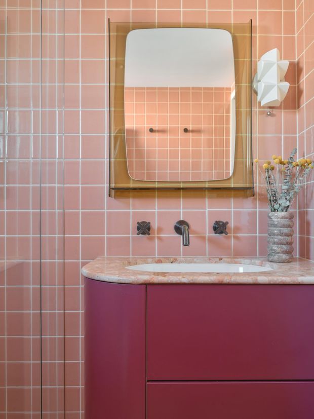 A compact, highly styled bathroom vanity area with pink grid-pattern ceramic wall tiles, a rounded rectangular mirror in a timber-toned frame, and a deep burgundy-pink floating vanity. The vanity has a curved pink stone benchtop with an integrated basin, dark metal wall-mounted tapware, and a sculptural white wall light. A textured vase with dried flowers adds a decorative accent. The composition is warm, playful, and retro-inspired.