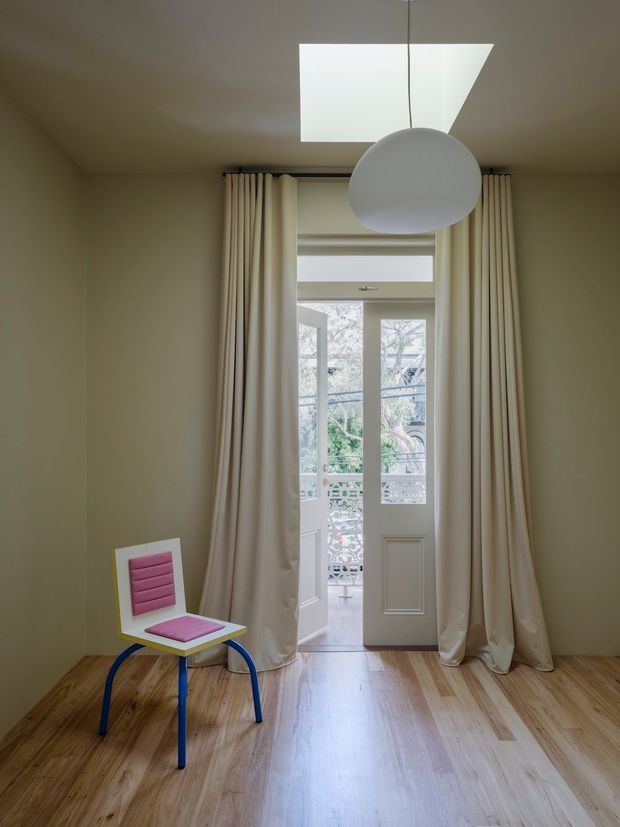 A compact, softly lit interior with pale beige walls, wide timber floorboards, full-height cream curtains and a white glazed French door opening to an exterior balcony or garden. A large white spherical pendant hangs beneath a rectangular skylight. A small brightly coloured chair with a pink seat and blue legs adds a playful accent.