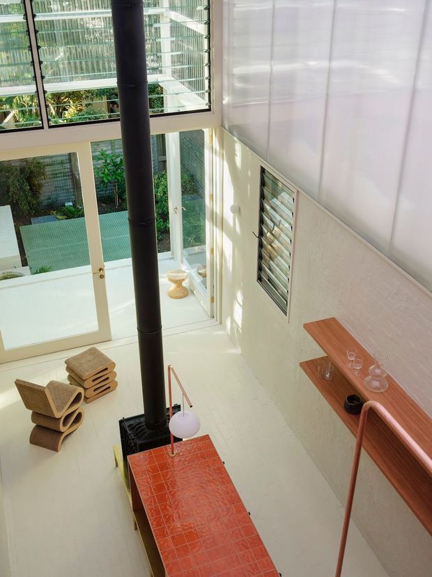 A tall, sunlit contemporary interior viewed from an elevated level, featuring a double-height glazed void with lush greenery visible outside. A black cylindrical flue rises from a red-orange tiled fireplace or stove enclosure, while warm timber shelving and a balustrade line the right side. Pale textured walls, large windows, louvred ventilation windows, and sculptural wooden seating create an airy, modern living space.