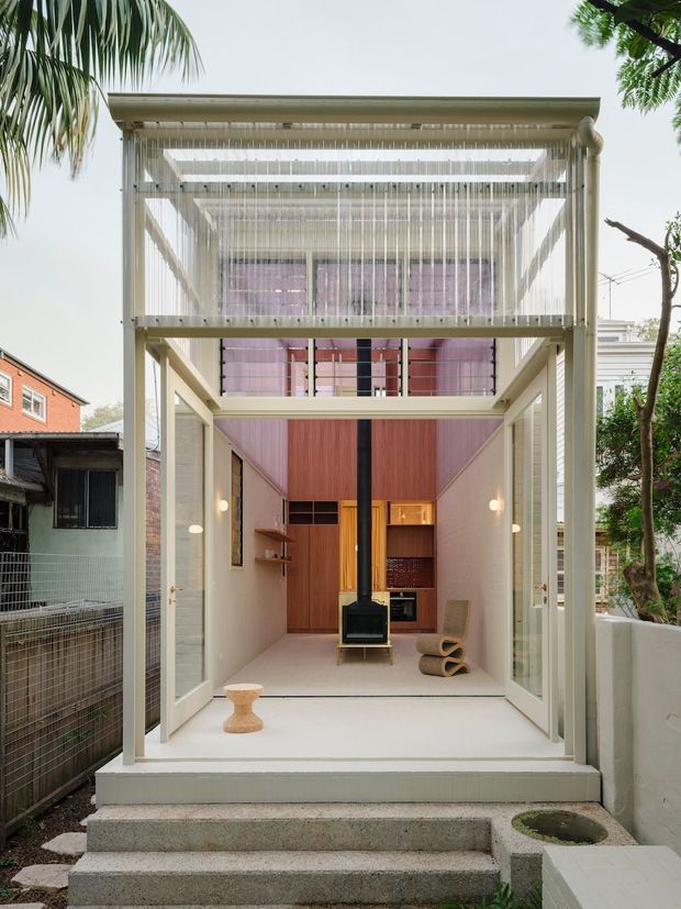A narrow, vertically oriented contemporary residential extension viewed from the garden. The open-fronted white structure has full-height glazed doors, a high pergola-like roof with hanging vertical screening, and a double-height interior. Inside are warm timber wall panels, a central black wood-burning stove and flue, built-in shelving, soft wall lighting, and a sculptural lounge chair. Concrete steps lead up to the light-filled space, with neighbouring urban homes and greenery around it.