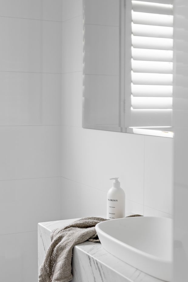 A minimalist white bathroom corner featuring large white wall tiles, a white vessel basin on a marble-look vanity, a frameless mirror, and white plantation shutters. A neutral taupe towel and soap dispenser add subtle contrast.