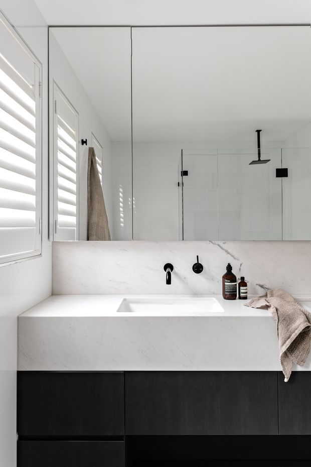 A minimalist contemporary bathroom vanity with a large frameless mirror, white marble-look wall and benchtop surfaces, integrated white basin, matte black tapware, and dark timber or black cabinetry. White plantation shutters and a glass shower enclosure are visible, creating a clean monochromatic scheme with warm grey towel and amber bathroom bottles as small accents.