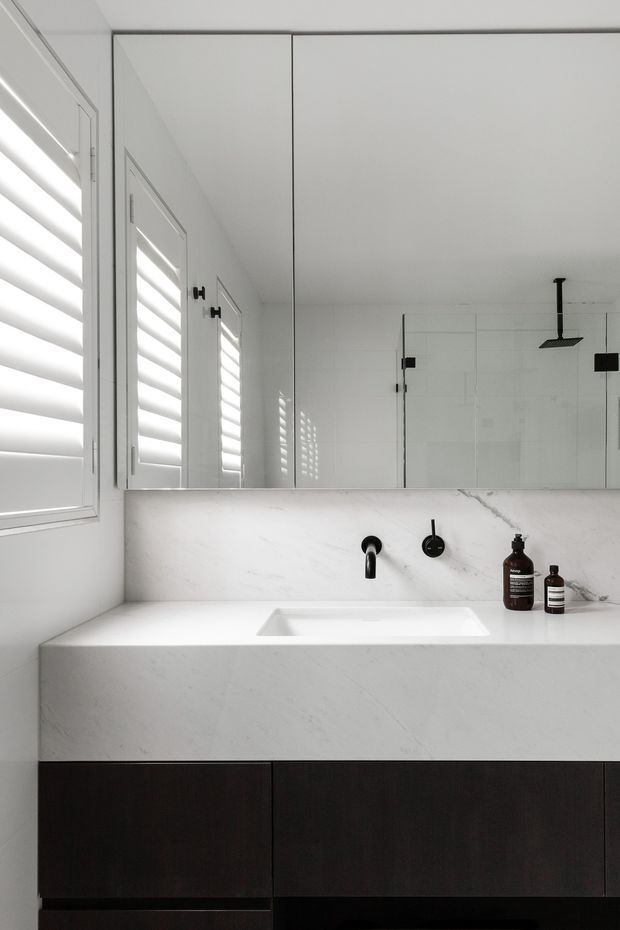 A bright contemporary bathroom vanity with a white veined stone benchtop and integrated rectangular basin, matte black wall-mounted tapware, dark timber cabinetry, a large mirrored cabinet, and white tiled walls. A frameless glass shower with a black ceiling-mounted showerhead is reflected in the mirror. Plantation shutters provide filtered daylight.