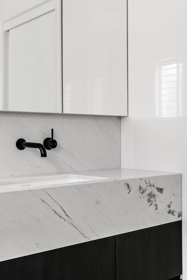 Close-up view of a refined bathroom vanity featuring a white marble-look benchtop and integrated basin, matching marble-look splashback, glossy white wall tiles, mirrored wall cabinetry, matte black wall-mounted tapware, and dark timber-look vanity cabinetry.
