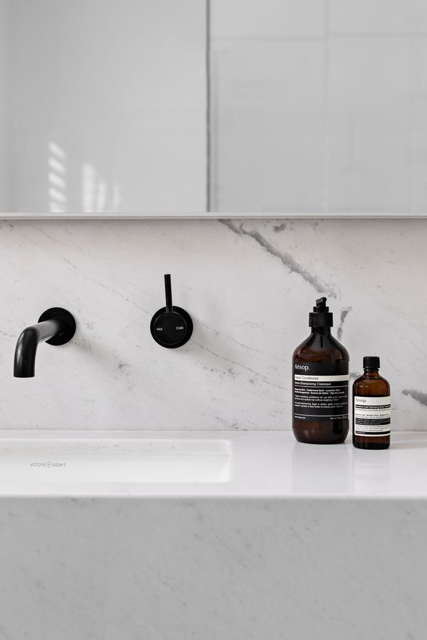 Close-up of a refined minimalist bathroom vanity featuring a white marble-look wall and countertop, integrated white basin, matte black wall-mounted faucet, and matching black temperature control. Amber bathroom product bottles sit on the counter, with a large mirror above.