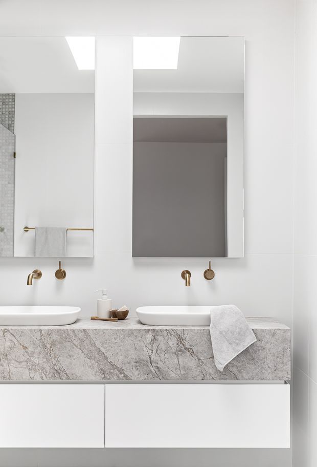 A bright contemporary double-vanity bathroom with two white vessel basins, wall-mounted brushed brass taps, large frameless mirrors, a pale grey marble-look stone benchtop, and streamlined white cabinetry. The space is predominantly white and grey with warm brass accents and minimal styling.