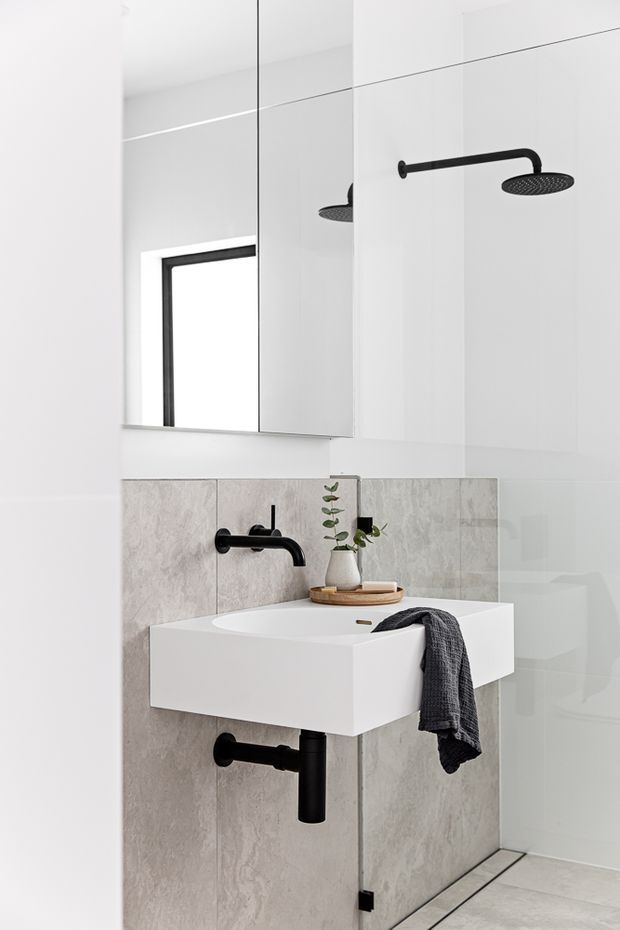 A bright, minimalist bathroom featuring a wall-mounted white rectangular basin with matte black wall-mounted tapware and exposed black waste pipe. Large grey stone-look wall tiles form the vanity backsplash, while a frameless glass shower enclosure with a black rain shower is visible to the right. A large mirrored cabinet or wall mirror spans the upper wall, reflecting the shower and a black-framed window. A small potted plant and folded dark towel add understated styling.