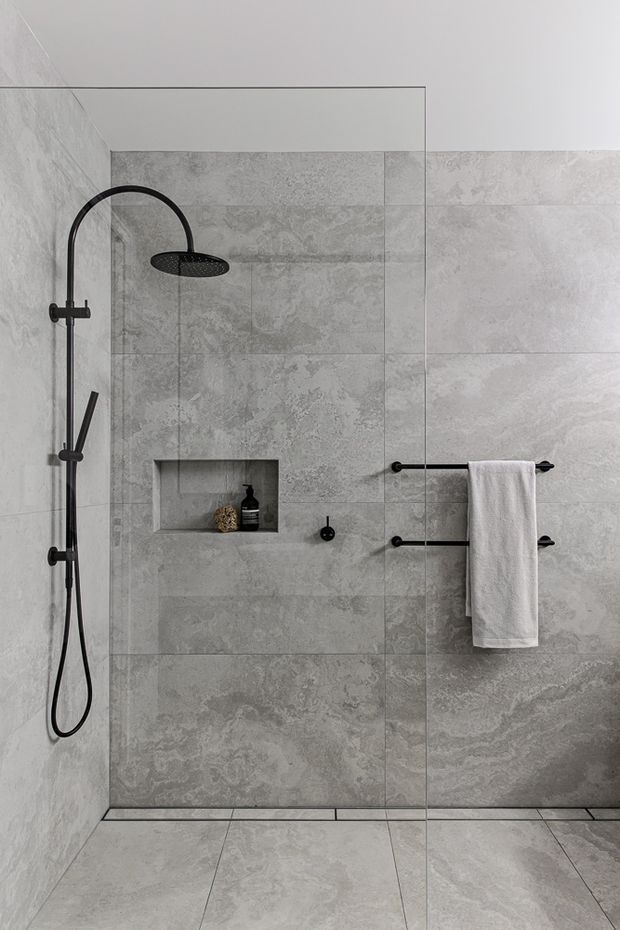 A minimalist walk-in shower enclosed by a frameless glass screen. Large-format grey stone-look tiles cover the walls and floor, with matte black shower fittings, recessed wall niche, and twin black towel rails holding a white towel.